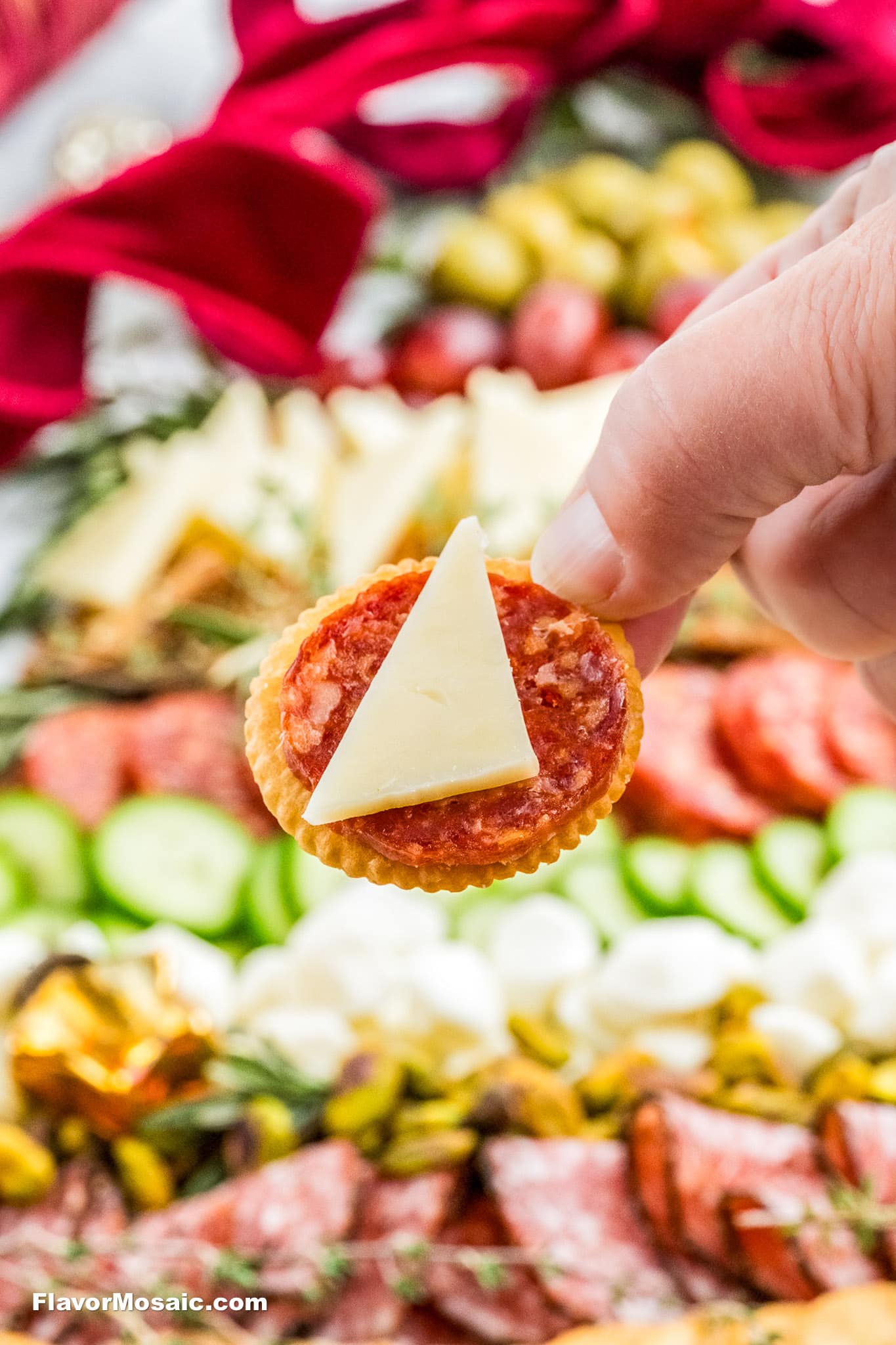 A hand holds a round cracker topped with a slice of salami and a triangle of cheese. In the blurred background, a colorful charcuterie board with cheese, meats, cucumbers, and other snacks is visible.