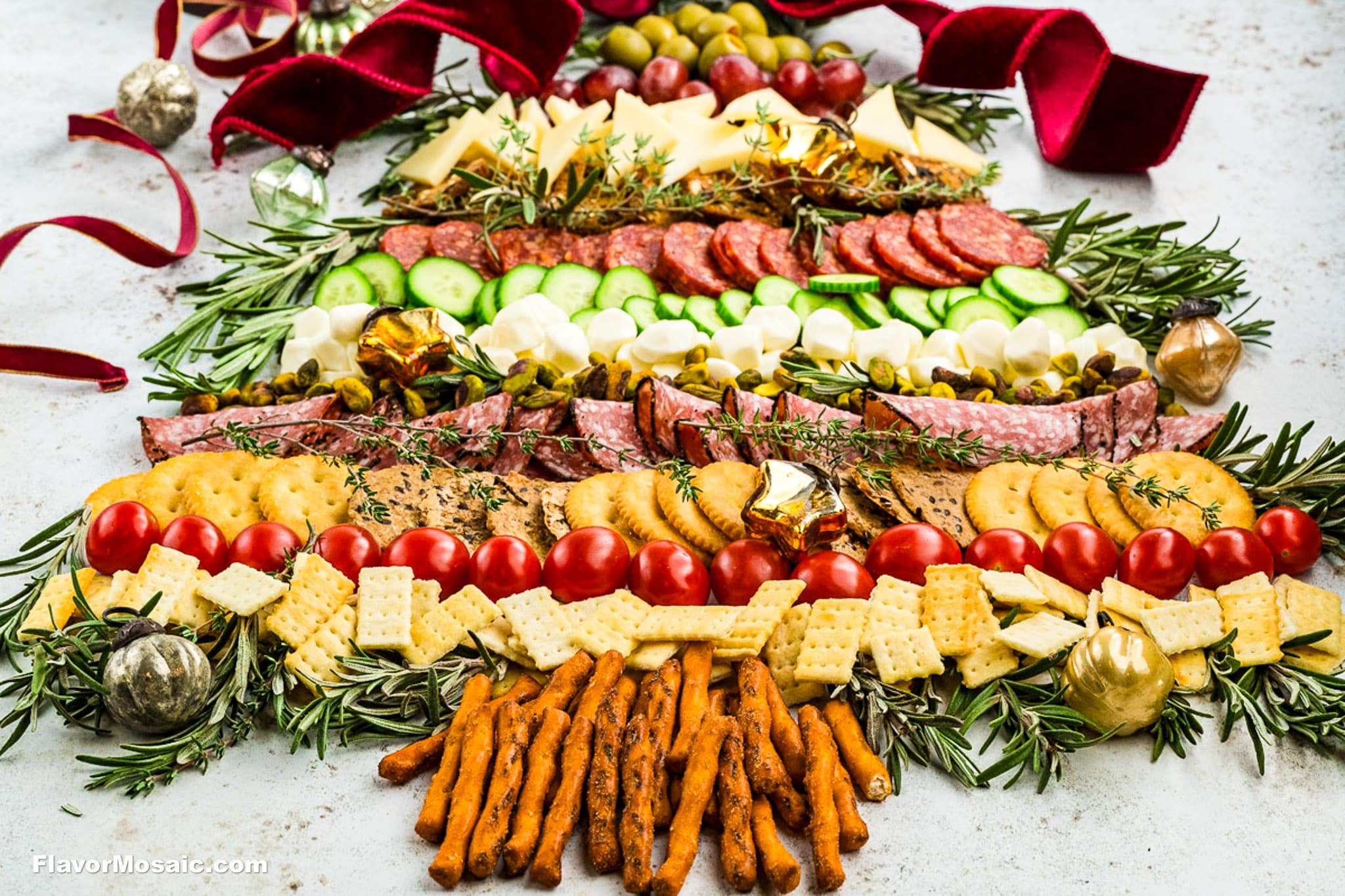 A festive Christmas Tree Charcuterie Board features crackers, pretzels, cheese, tomatoes, salami, cucumbers, olives, and herbs, all adorned with gold ornaments and a red ribbon for a merry holiday display.