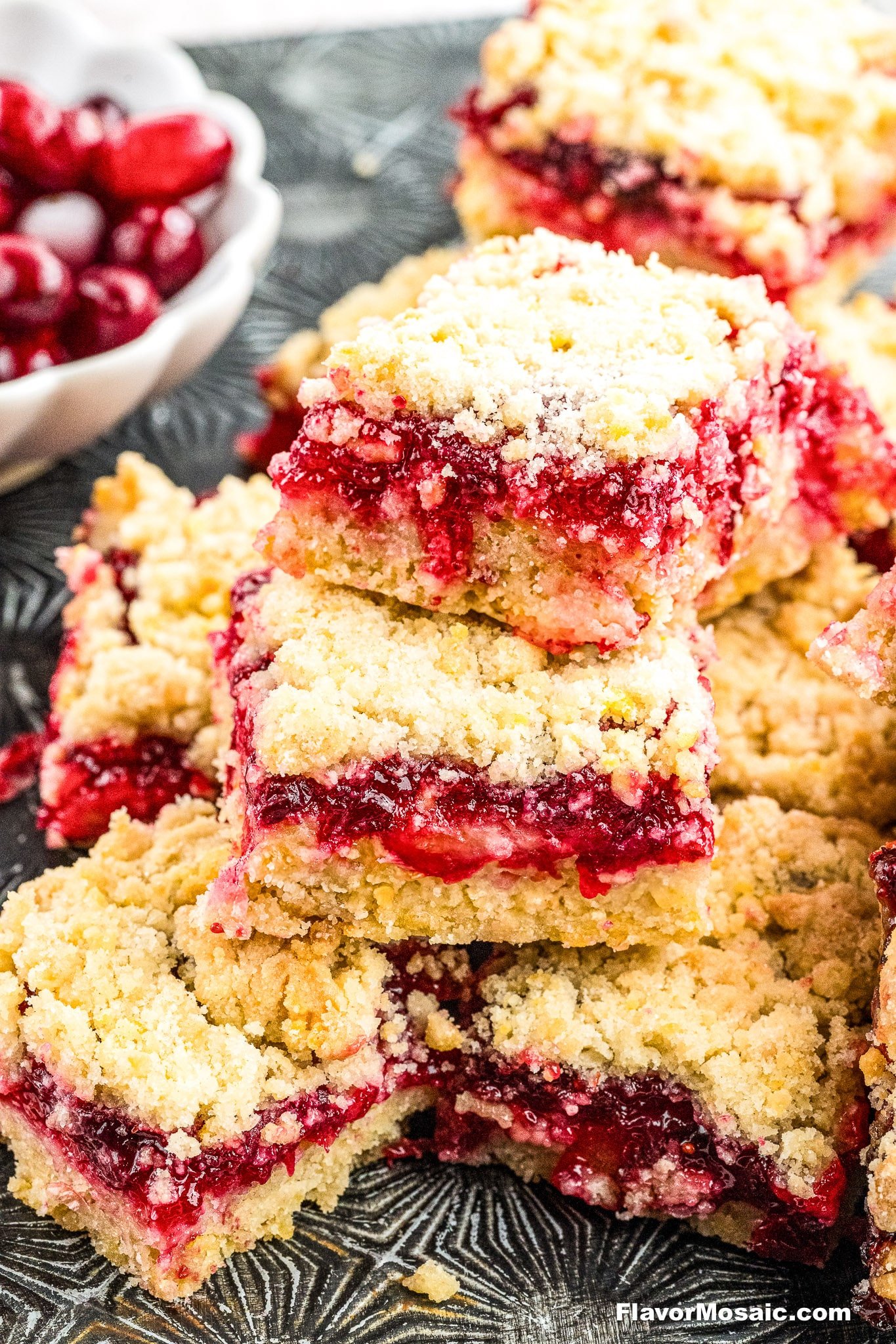 A stack of crumbly Cranberry Crumb Bars with a vibrant red berry filling sits on a patterned tray. A bowl of fresh cranberries is visible in the background.
