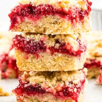 Three crumbly dessert bars with golden crusts and vibrant red fruit filling, possibly raspberry, are stacked on top of each other, with more bars blurred in the background.