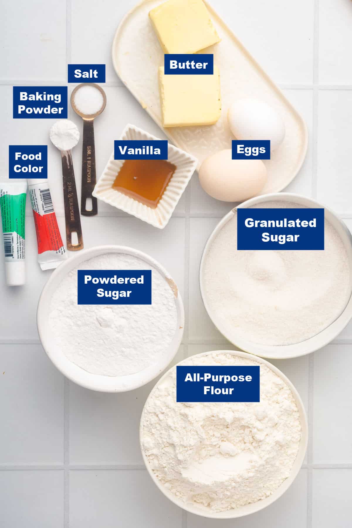 Ingredients for baking are arranged on a white surface: butter, eggs, granulated sugar, powdered sugar, all-purpose flour, vanilla, salt, baking powder, and food coloring tubes. Each item is labeled.