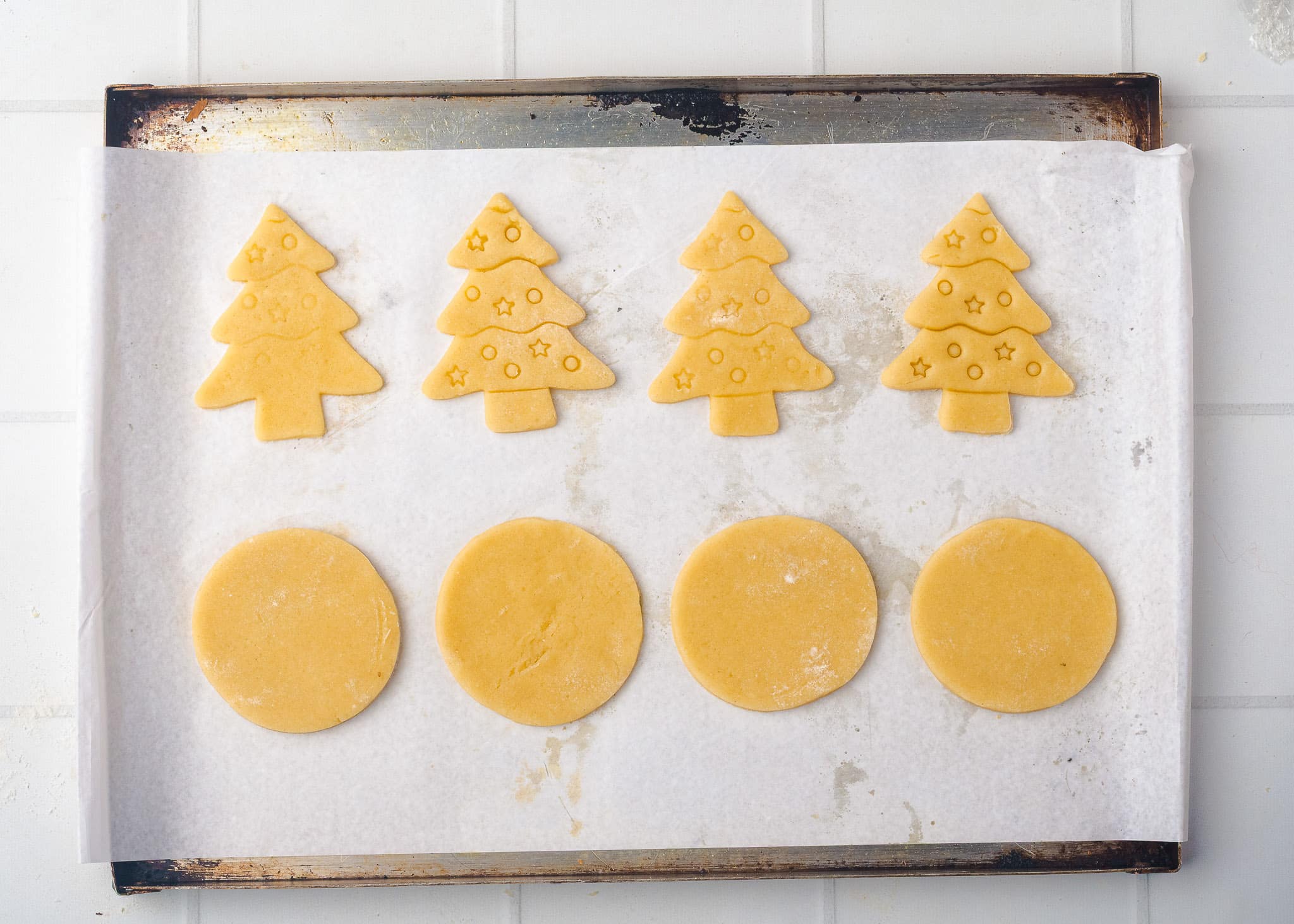 A baking sheet lined with parchment paper displays six unbaked sugar cookies: three round ones and three Christmas tree-shaped ones, decorated with small star imprints. The sheet rests on a white tiled surface.