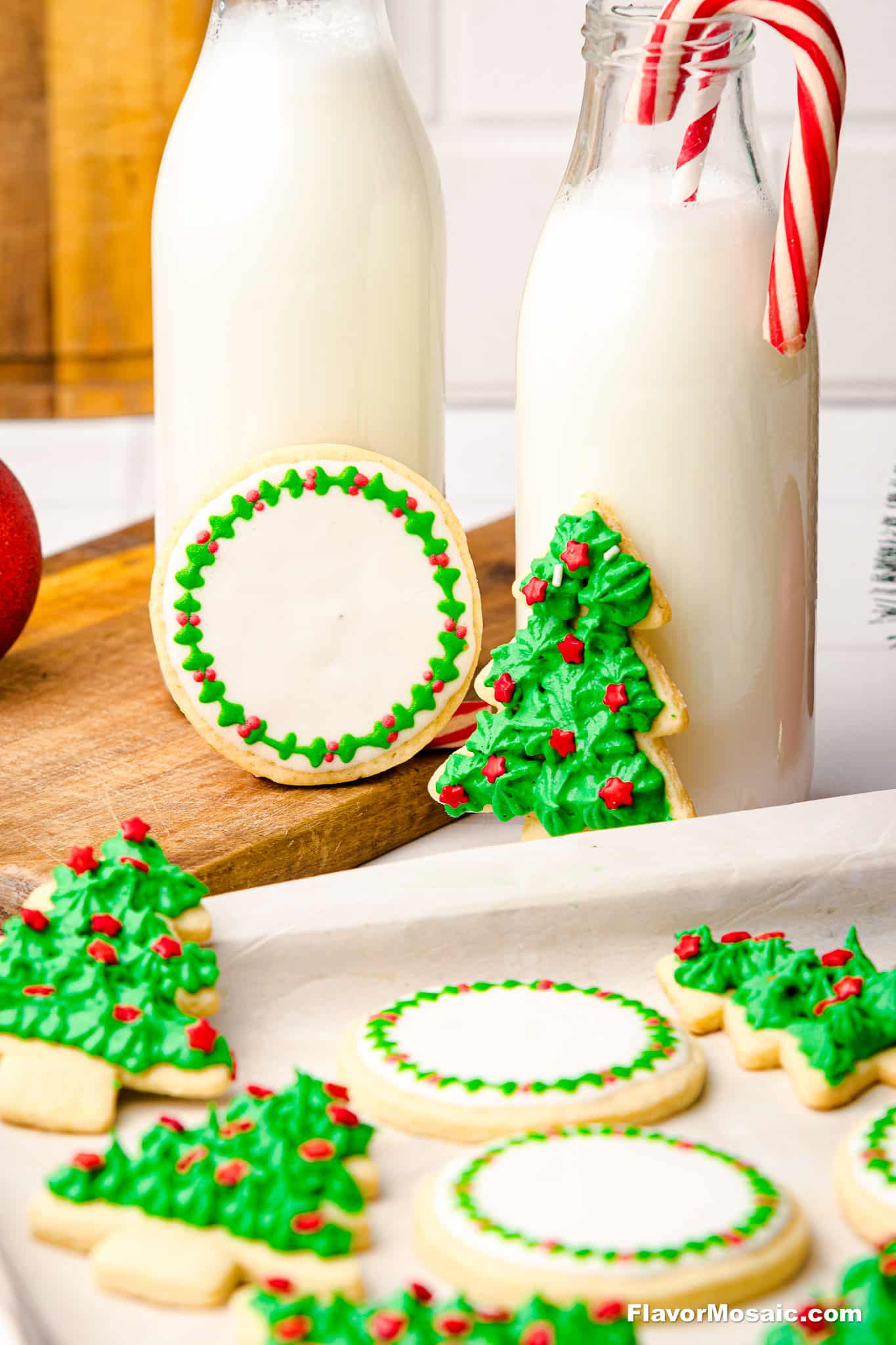 Sugar cookies decorated as Christmas trees and wreaths with green and red icing are displayed on a table next to two bottles of milk, one with a candy cane inside.
