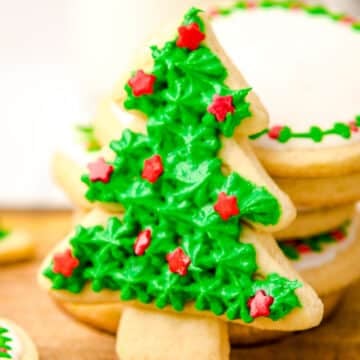 A Christmas tree-shaped sugar cookie decorated with green icing and red star-shaped sprinkles is propped against a stack of round, iced sugar cookies on a wooden surface.