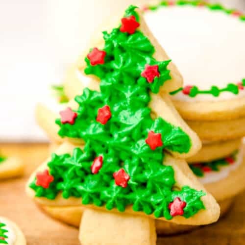 A Christmas tree-shaped sugar cookie decorated with green icing and red star-shaped sprinkles is propped against a stack of round, iced sugar cookies on a wooden surface.