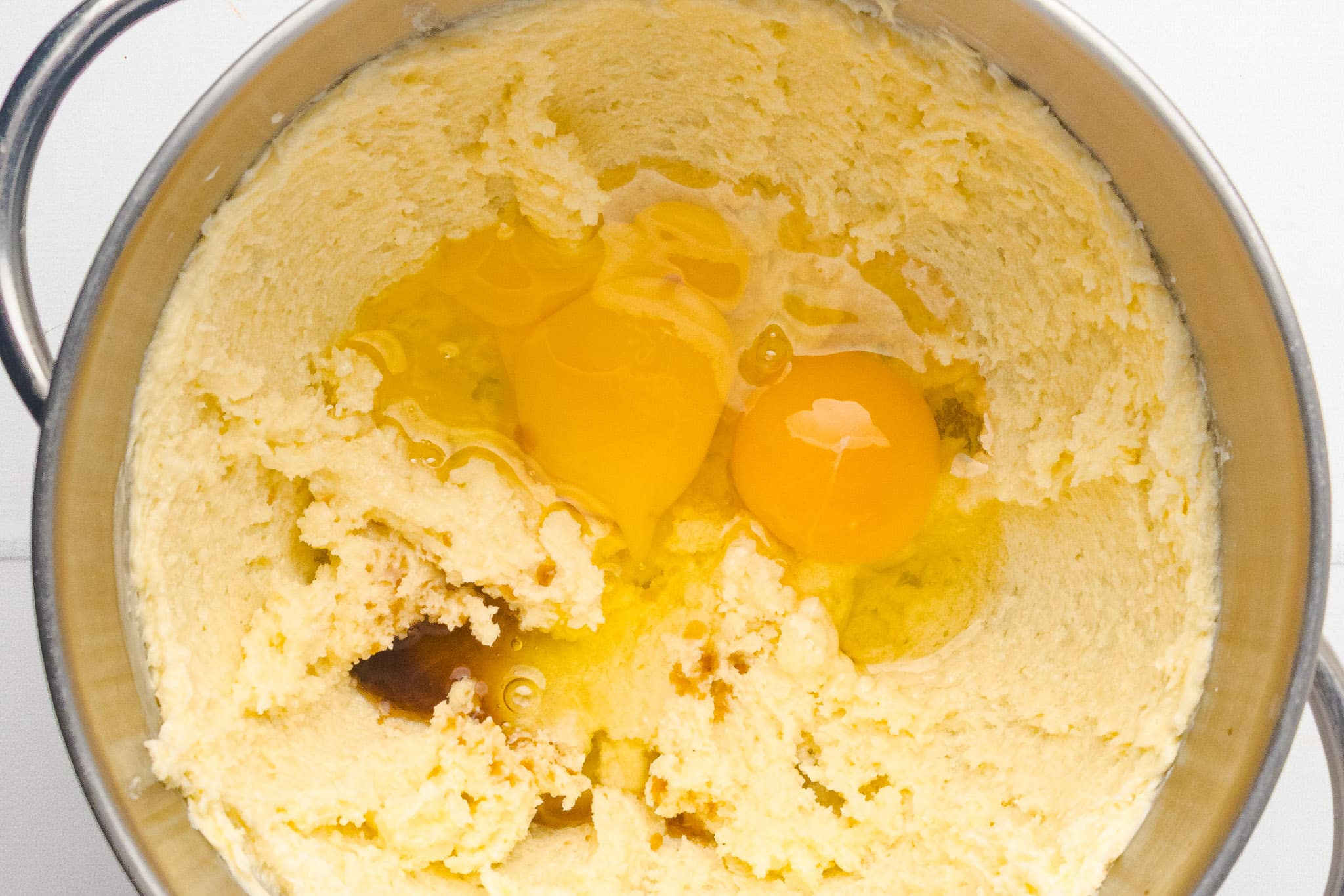 A metal mixing bowl containing creamed butter and sugar with two cracked eggs and a splash of vanilla extract on top, ready to be mixed for baking.