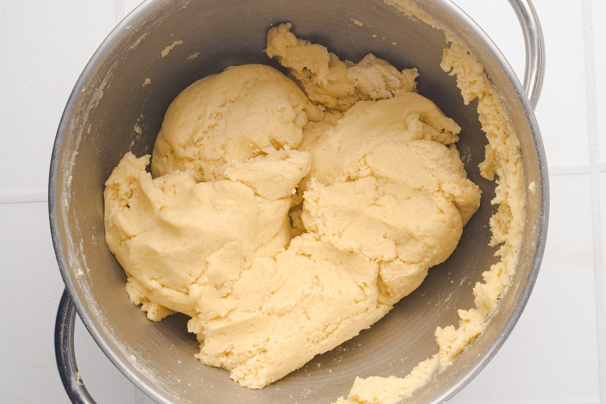 A metal mixing bowl filled with thick, pale yellow dough, some of which is sticking to the sides of the bowl. The bowl is placed on a white tiled surface.