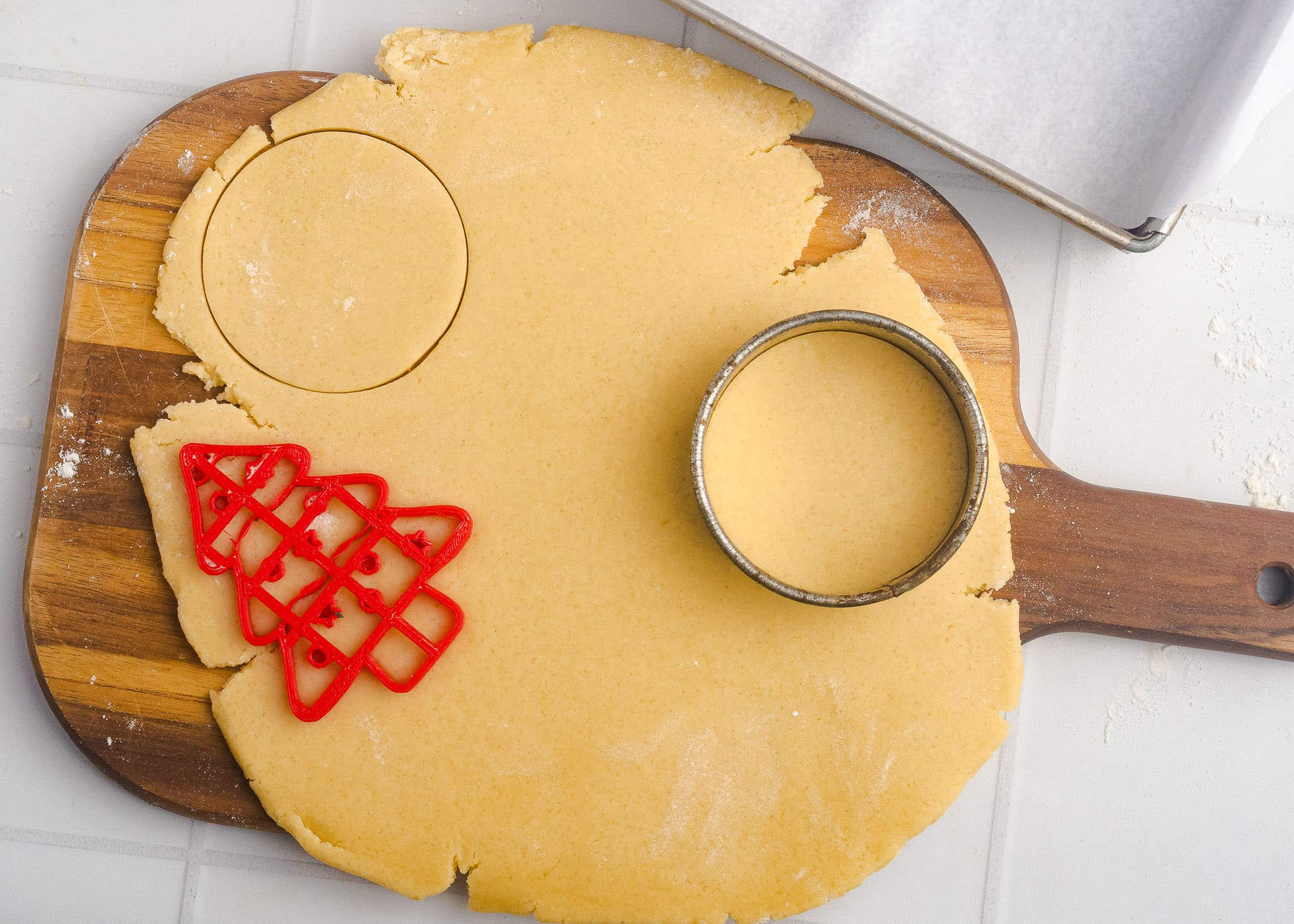 A wooden board with rolled-out cookie dough. A round cookie cutter and a red Christmas tree-shaped cutter are pressing shapes into the dough. A baking tray lined with parchment paper is nearby.