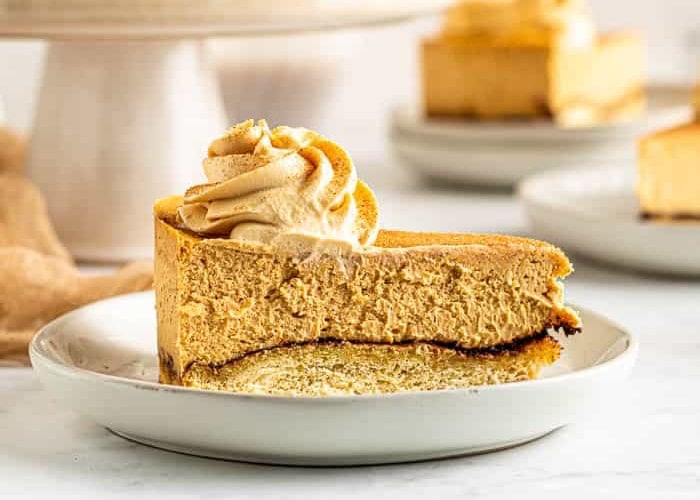 A slice of creamy pumpkin cheesecake with a swirl of whipped topping sits on a white plate, with more cheesecake in the blurred background.