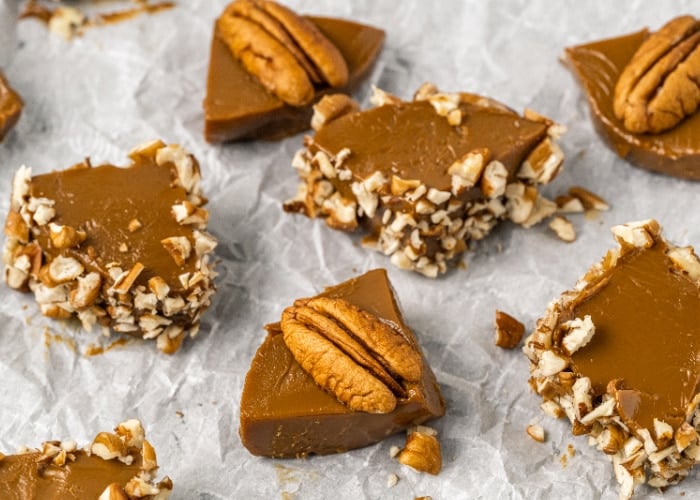 Pieces of caramel candy topped with pecans and coated on the sides with chopped nuts are arranged on crumpled parchment paper.