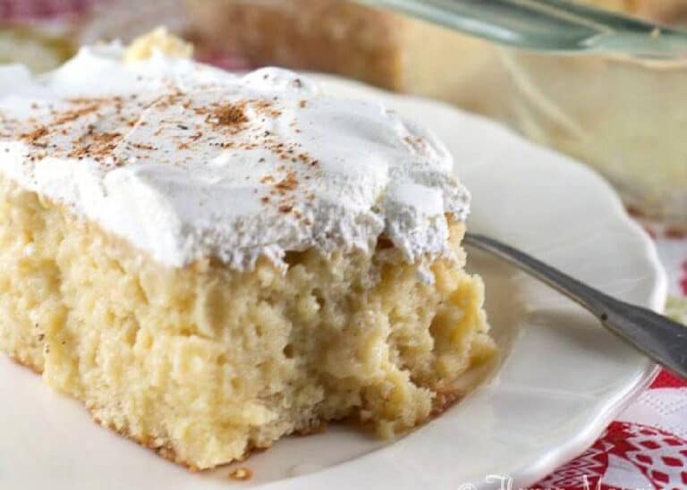 A slice of moist tres leches cake topped with whipped cream and a sprinkle of cinnamon, served on a white plate with a fork.