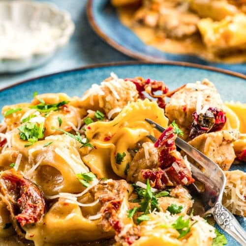 A plate of creamy tortellini pasta with sun-dried tomatoes, pieces of chicken, and parmesan cheese, garnished with chopped parsley, served on a blue ceramic plate with a fork.