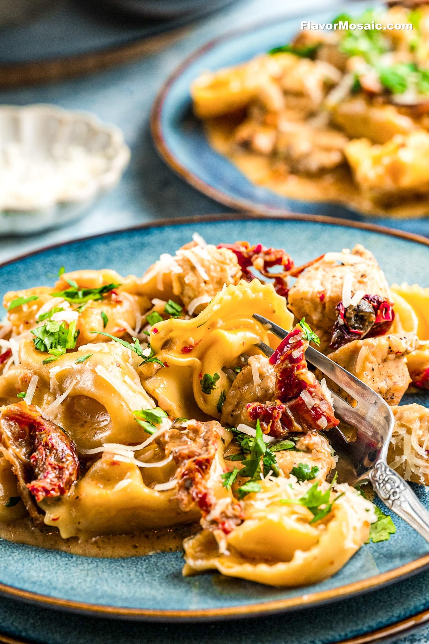 A plate of creamy tortellini pasta with sun-dried tomatoes, pieces of chicken, and parmesan cheese, garnished with chopped parsley, served on a blue ceramic plate with a fork.