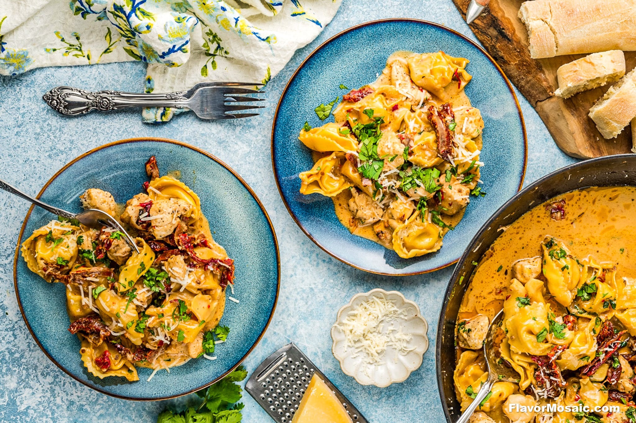 Overhead view of two blue plates with creamy tortellini pasta, chicken, and sun-dried tomatoes, garnished with herbs. A skillet of the same dish, grated cheese, and bread are nearby on a light blue table.