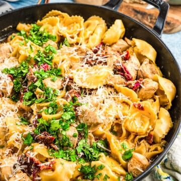 A skillet filled with cheesy pasta, chunks of chicken, sun-dried tomatoes, and fresh parsley, topped with grated parmesan. In the background, slices of bread are on a wooden board.