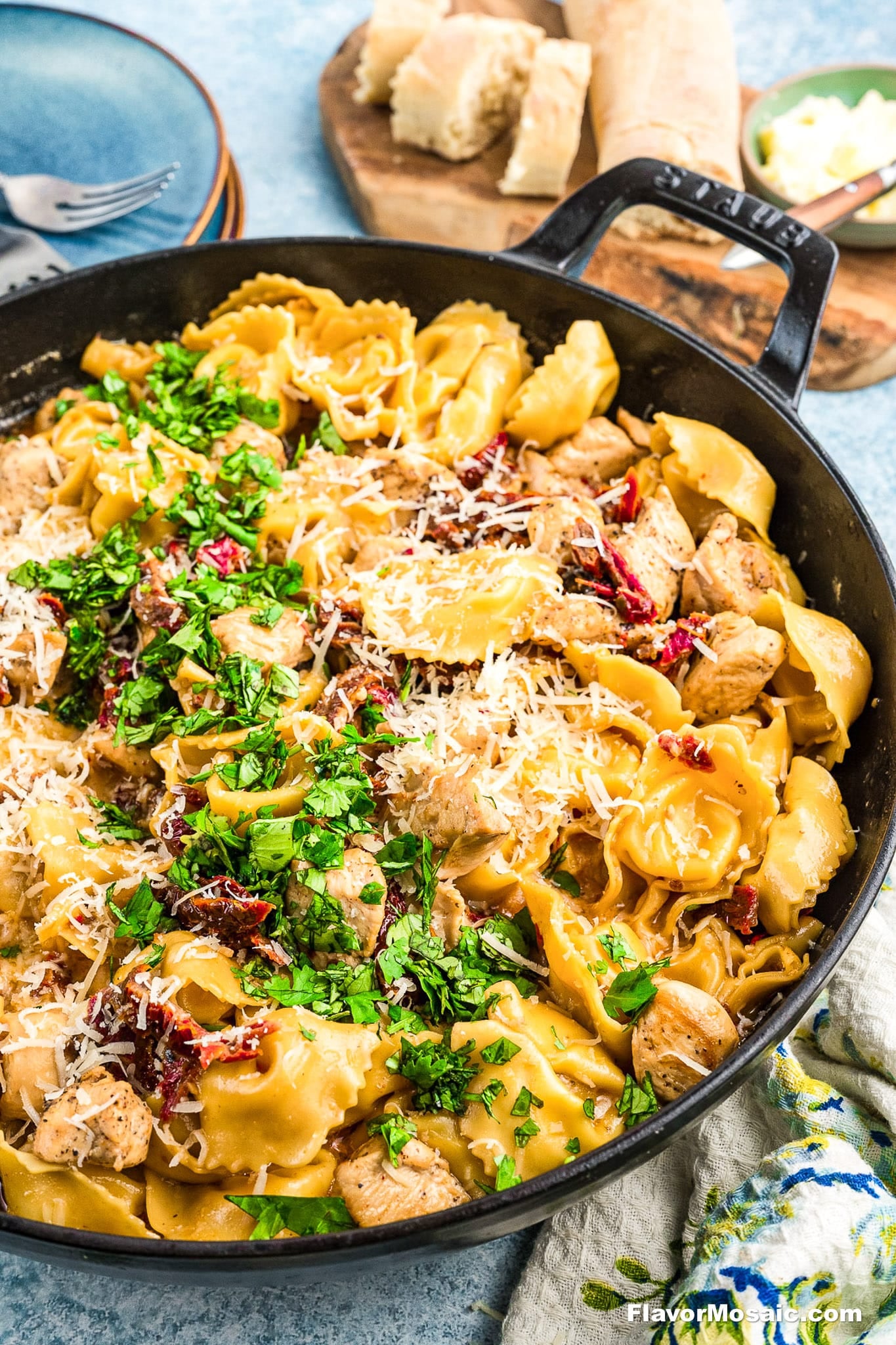 A skillet filled with cheesy pasta, chunks of chicken, sun-dried tomatoes, and fresh parsley, topped with grated parmesan. In the background, slices of bread are on a wooden board.