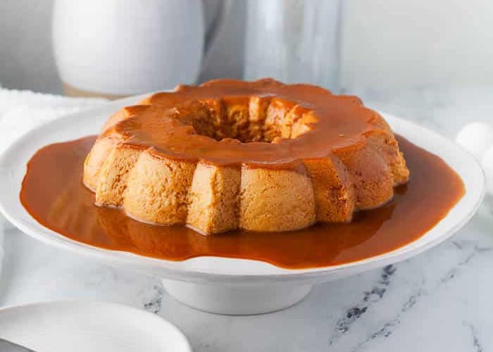 A round caramel flan sits on a white cake stand, covered in smooth caramel sauce that drips down the sides, with a marble surface and blurred kitchen items in the background.
