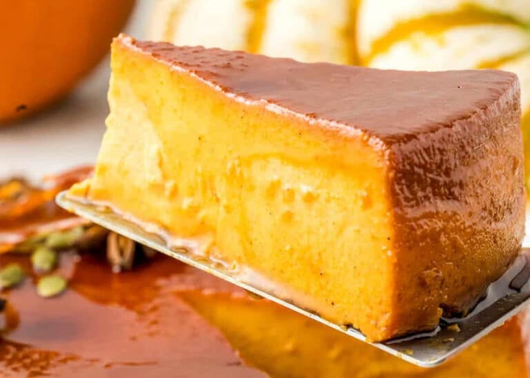 A close-up of a creamy slice of flan with a shiny caramel topping on a spatula, with more flan and blurred pumpkins in the background.