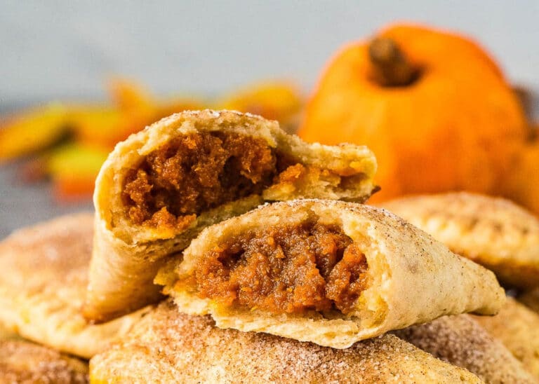 A close-up of pumpkin hand pies, one split open to reveal a spiced orange pumpkin filling. More pies and a small pumpkin are blurred in the background.