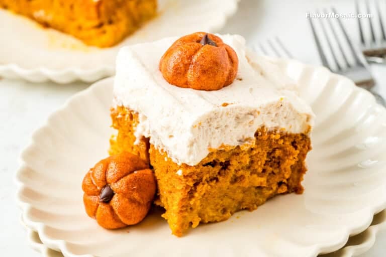 A slice of pumpkin cake with white frosting sits on a white plate, garnished with two small pumpkin-shaped candies. A fork and another plate with cake are visible in the background.