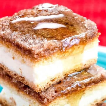 Two stacked cheesecake bars with a cinnamon-sugar topping and a drizzle of syrup, served on a turquoise plate with a red background.