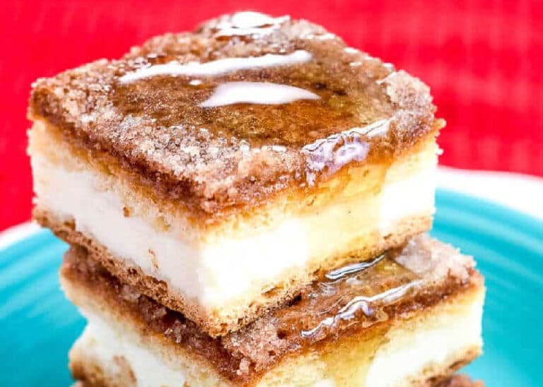 Two stacked cheesecake bars with a cinnamon-sugar topping and a drizzle of syrup, served on a turquoise plate with a red background.