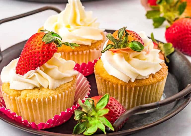 Three vanilla cupcakes with swirls of white frosting, each topped with a fresh strawberry, are arranged on a round metal tray with extra strawberries scattered around them.
