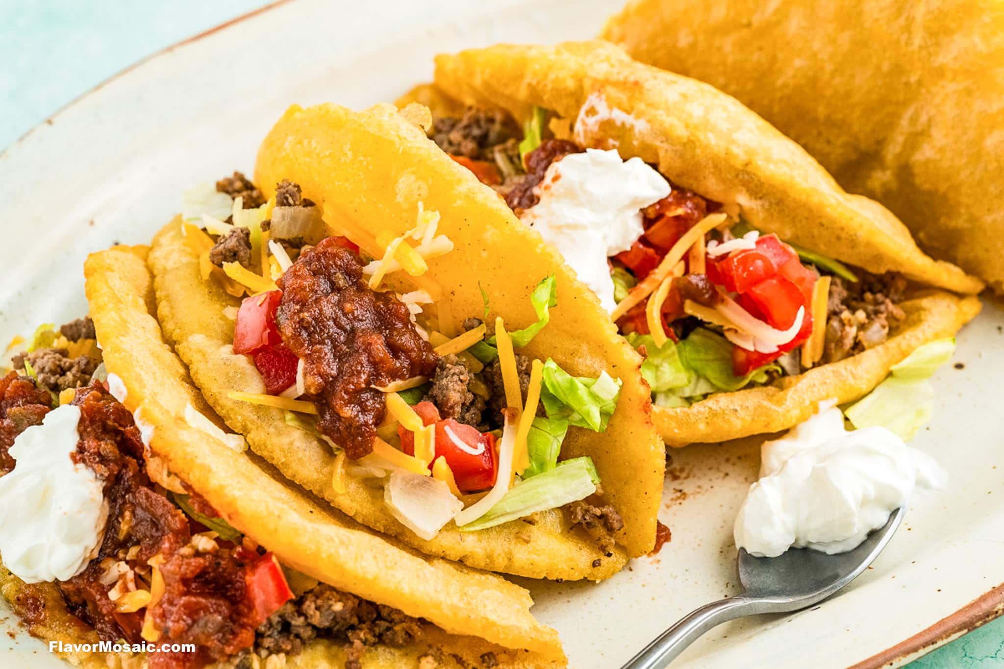 Three crispy tacos filled with seasoned ground beef, shredded lettuce, diced tomatoes, cheddar cheese, salsa, and sour cream are arranged on a plate, with an extra dollop of sour cream on a spoon nearby.