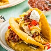 Two crispy tacos filled with ground beef, shredded lettuce, diced tomatoes, shredded cheese, salsa, and a dollop of sour cream on a white plate, with salsa and more food in the background.