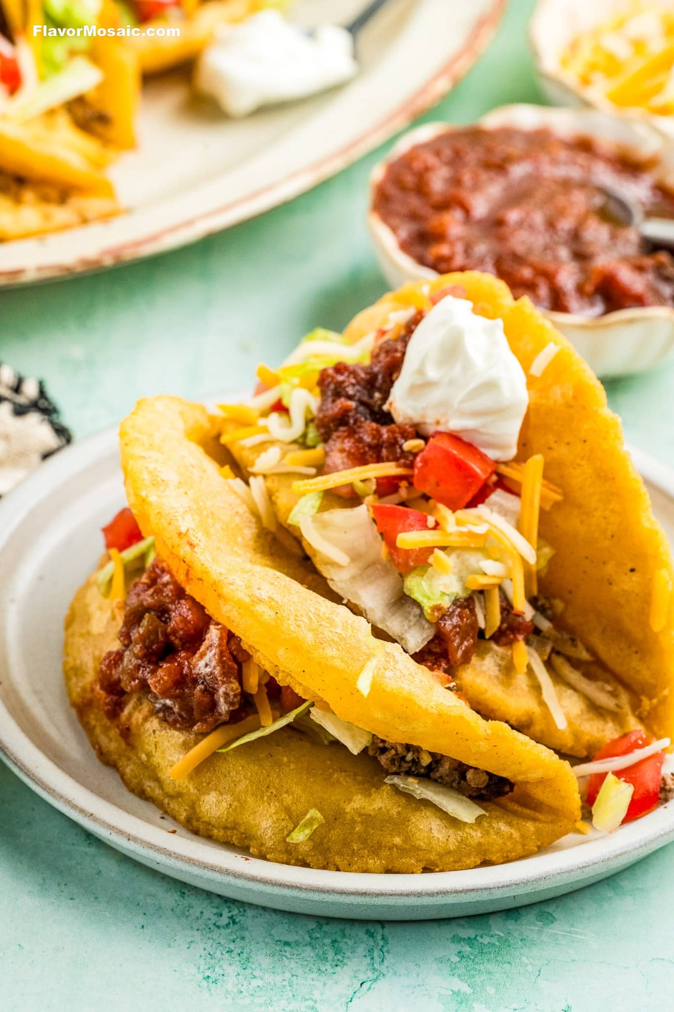 Two crispy tacos filled with ground beef, shredded lettuce, diced tomatoes, shredded cheese, salsa, and a dollop of sour cream on a white plate, with salsa and more food in the background.