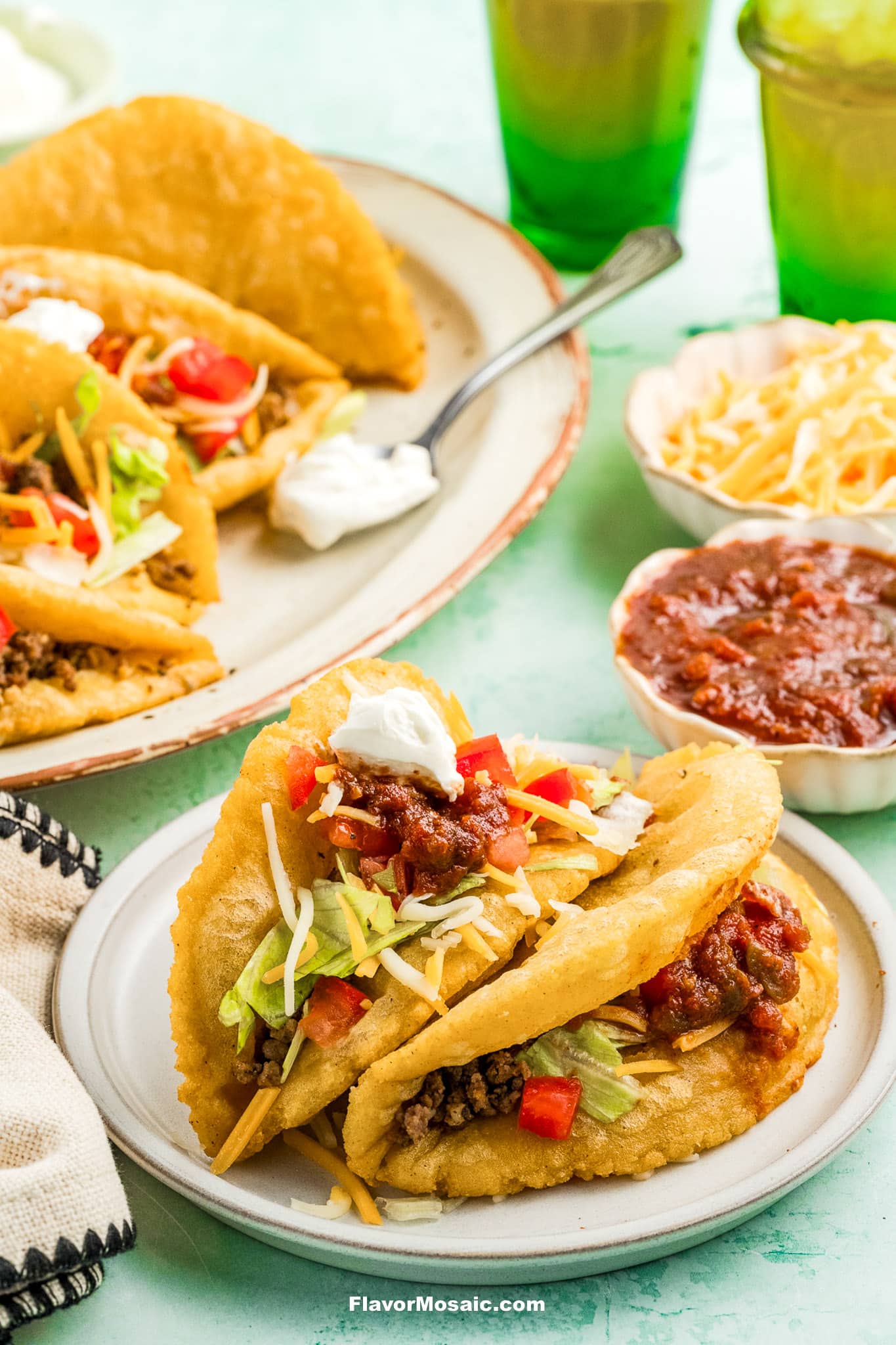 Two crispy tacos filled with ground beef, lettuce, shredded cheese, diced tomatoes, salsa, and a dollop of sour cream on a plate. Additional tacos, salsa, shredded cheese, and drinks are in the background.