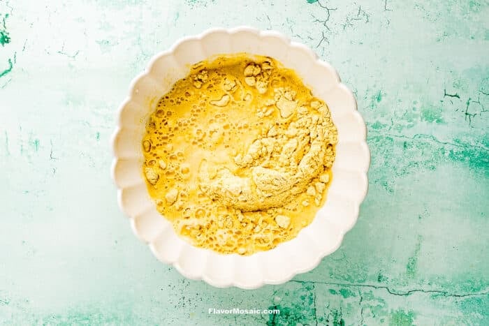 A white scalloped bowl containing a mixture of dry yellow ingredients and liquid, partially blended, sits on a textured light green surface.