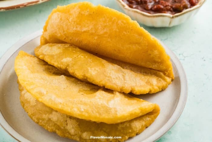 Three golden, crispy empanadas are arranged on a white plate, with a bowl of salsa visible in the background. The empanadas have a lightly fried texture and folded shape.