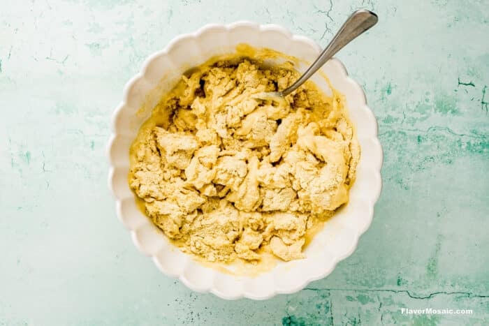 A white scalloped bowl filled with thick, yellowish dough mixture and a metal spoon, set on a light blue textured surface.