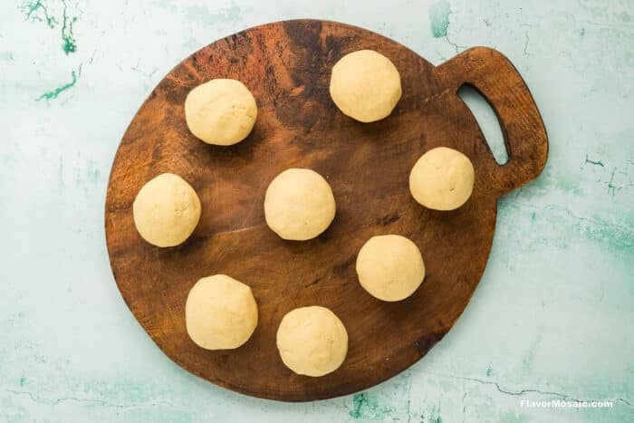 Nine evenly spaced balls of dough sit on a round wooden cutting board with a handle, set against a light turquoise textured background.