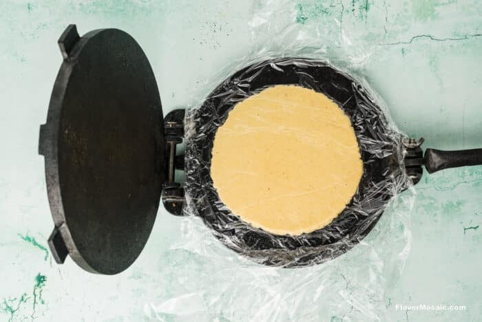 A round tortilla dough sits between two sheets of plastic wrap on an open cast iron tortilla press, placed on a light green textured surface.