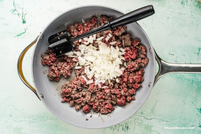 Ground beef and chopped onions cooking in a gray skillet, with a black spatula resting on the side, on a light green textured background.