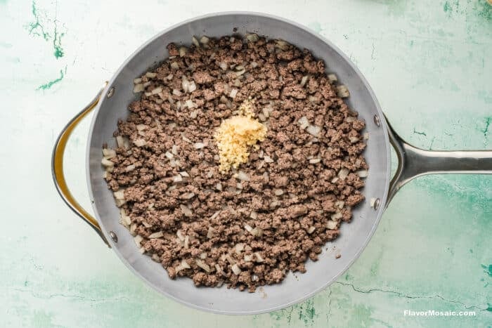 A skillet containing cooked ground beef and chopped onions, with a small mound of minced garlic in the center, sits on a light green marbled surface.