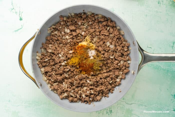 A skillet filled with cooked ground beef, chopped onions, and various colorful spices in the center, ready to be mixed together. The pan is resting on a light green textured surface.