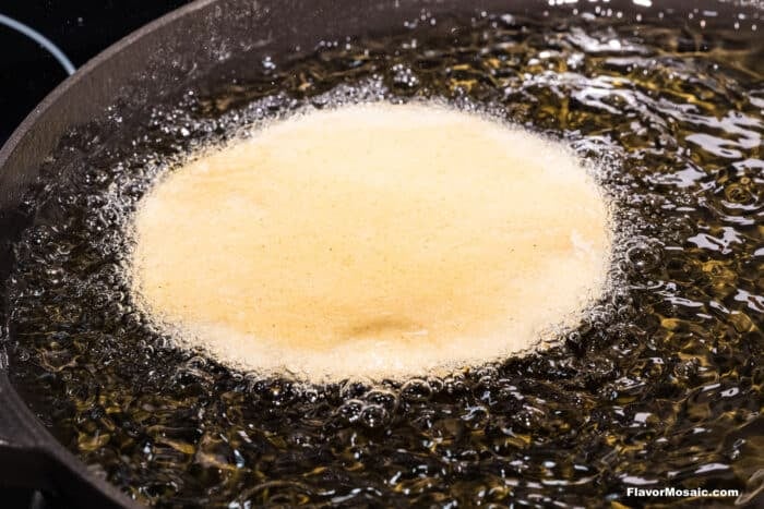 A round tortilla is frying in hot oil in a black skillet, with bubbles forming around its edges as it cooks.
