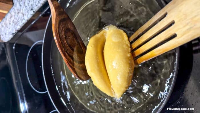 A pair of dumplings being deep-fried in hot oil, held by a wooden spoon and a wooden fork over a stovetop.