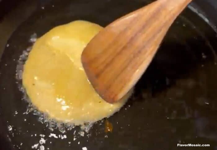 A wooden spatula presses a round, golden batter in a skillet, frying in hot oil with visible bubbles forming at the edges.