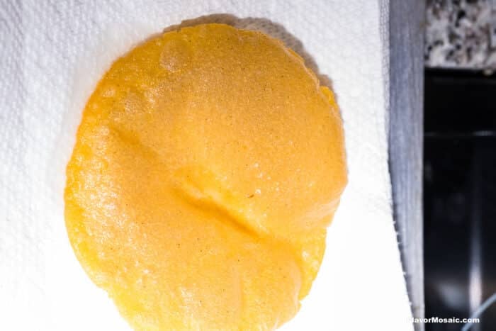 A close-up of a freshly cooked, golden brown taco shell resting on a white paper towel, with part of a granite countertop and stove visible in the background.