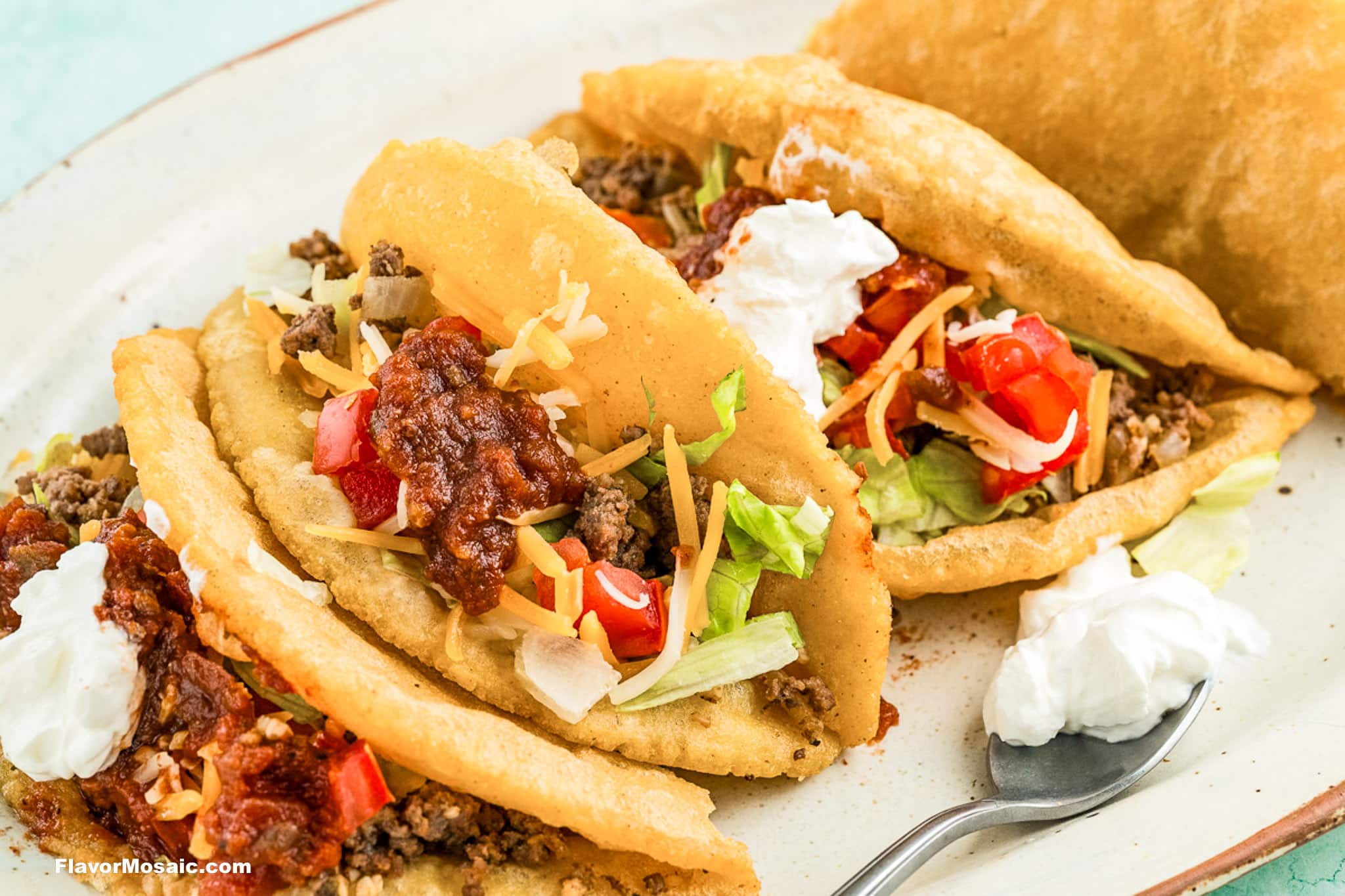 Three crispy tacos filled with ground beef, shredded cheese, lettuce, diced tomatoes, salsa, and sour cream are served on a white plate, with a spoonful of sour cream on the side.