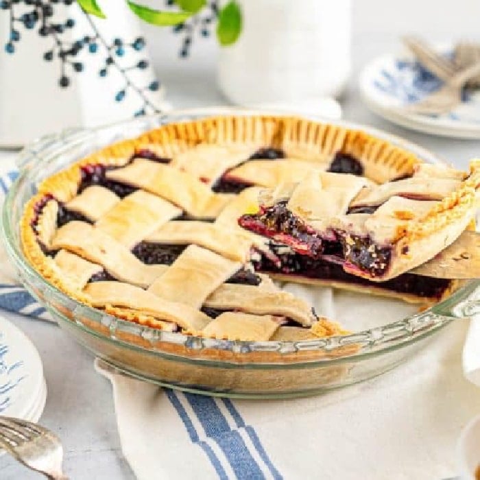A baked blackberry pie with a lattice crust in a glass pie dish. One slice is being served with a pie server. Plates and utensils are visible in the background on a white and blue tablecloth.