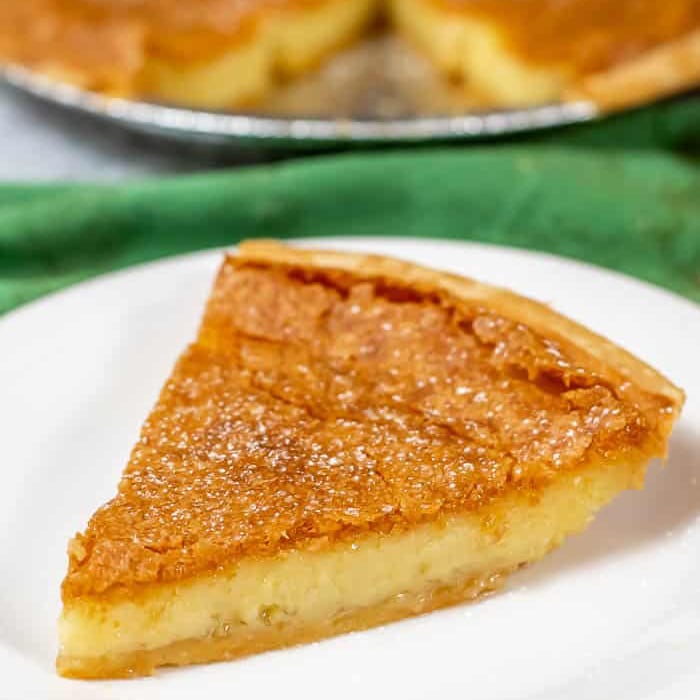 A close-up of a single slice of chess pie on a white plate, showing a golden-brown, slightly crackled top and creamy filling, with the rest of the pie visible in the background.