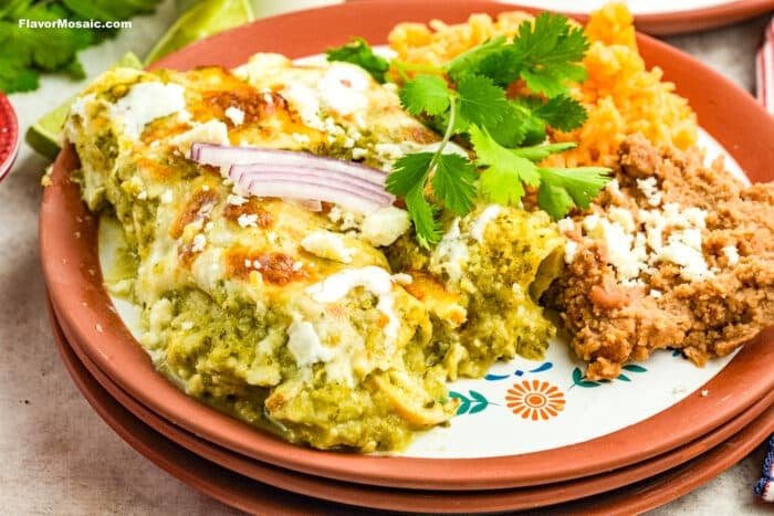 A plate of green enchiladas topped with sour cream, red onion, and cilantro, served with Mexican rice and refried beans garnished with cheese on a decorative plate.