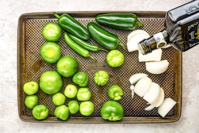 A baking sheet with fresh tomatillos, jalapeños, serrano peppers, chopped white onion, and olive oil being poured over the vegetables, ready for roasting.