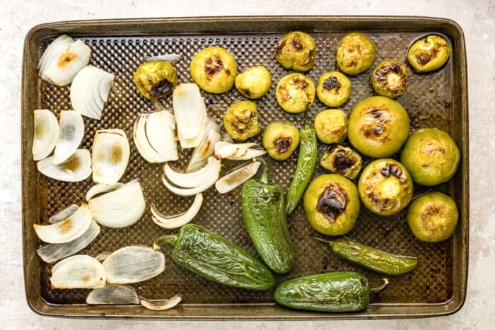 A baking sheet with roasted tomatillos, jalapeños, and onion slices, showing charred spots and caramelized edges on the vegetables.