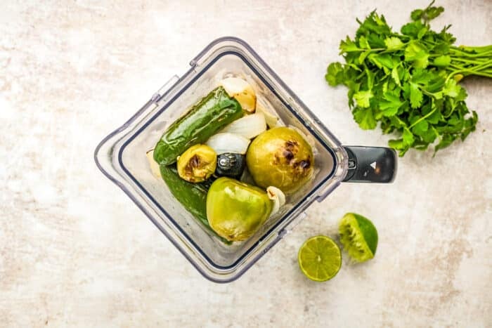 A blender filled with green vegetables and ingredients, including tomatillos, jalapeños, onion, and garlic. Next to the blender are fresh cilantro and a halved lime on a light-colored surface.